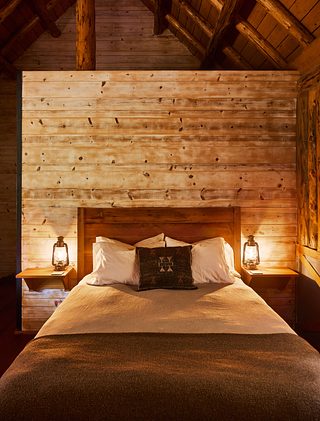 A warmly lit cabin bedroom with a wooden bed, white pillows, lanterns on floating nightstands, and exposed log beams overhead.