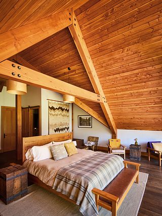Rustic guest room with a king bed under a dramatic vaulted wood ceiling, exposed timber beams, warm wood floors, woven wall art, and cozy seating arranged around the room.