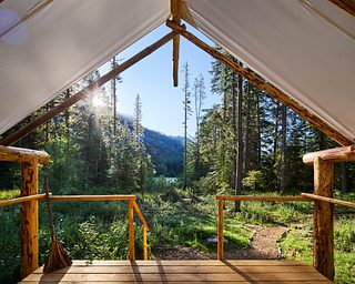 View from inside a canvas tent, looking past a covered wooden porch into a sunlit evergreen forest with mountain slopes visible through the trees.