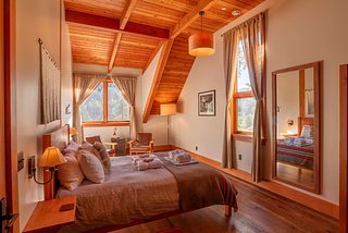 Warm, rustic guest room with a bed under a vaulted wood ceiling with exposed beams, tall curtained windows overlooking trees, wood floors, wall sconces, a full-length mirror, and a small sitting area by the window.