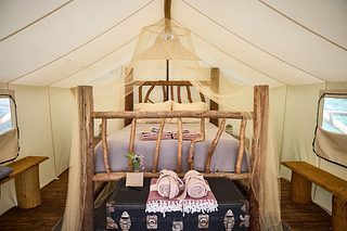 A cozy canvas tent interior with a rustic wood-frame bed under a draped mosquito net, folded towels at the foot, a trunk and flowers in front, and simple wooden benches along both sides.