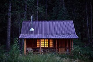 A log cabin glows from within at dusk, set among dense forest trees and tall grass.