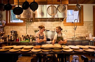 Two chefs talk in a warm, rustic commercial kitchen, standing behind a counter lined with plates and a tray of prepared fish, with pots and strainers hanging overhead.