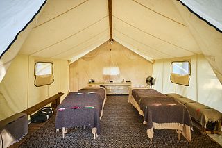A spacious canvas tent set up as a massage room, with two massage tables covered in blankets, small side benches, a patterned rug, and soft daylight filtering through the tent walls.