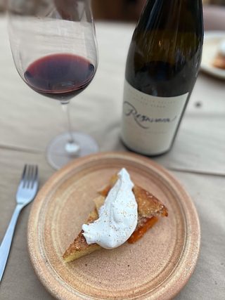 A slice of a baked dessert topped with whipped mascarpone and a dusting of nutmeg next to a bottle & glass of red wine.
