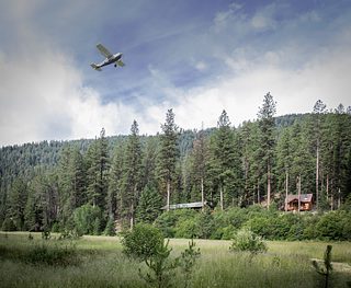 A small airplane flies low over a forested meadow, with tall pine trees, rolling hills, and rustic cabins visible below.