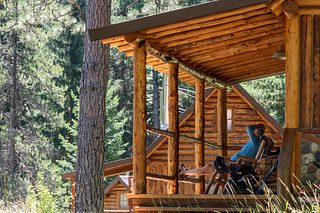 A person reclines in a chair on the porch of a rustic log cabin, resting in the shade among tall trees and meadow grass.