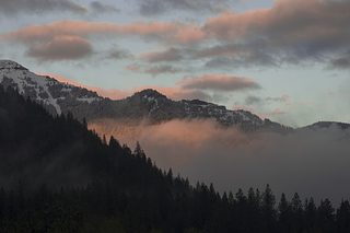 Low mist drifts through dark mountain ridges at sunset, with pink clouds and soft light catching the higher slopes.