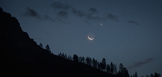 A thin crescent moon and a bright planet shine in the deep blue evening sky above the dark silhouette of a mountain ridge and treetops.