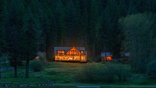 A large lodge glows warmly from within at the edge of a dark forest clearing, with nearby cabins faintly visible among the trees.