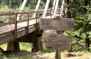 A weathered wooden trail sign stands beside a bridge, pointing toward Horse Ranch, Moss Spring, and Minam River trails at the edge of a forest.
