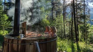 Two women relax in a steaming outdoor wooden hot tub set among trees, with forest and mountain views in the background.