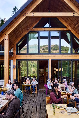 Guests dine on the wooden deck of a lodge with a large A-frame roof and tall glass windows reflecting the surrounding mountains and forest.