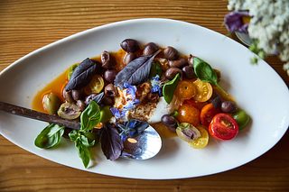 A colorful plated dish of olives, cherry tomatoes, basil leaves, and edible flowers arranged around a creamy center.