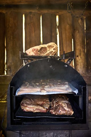 Large cuts of meat smoke on racks inside a black smoker, with smoke rising against a rustic wooden backdrop.