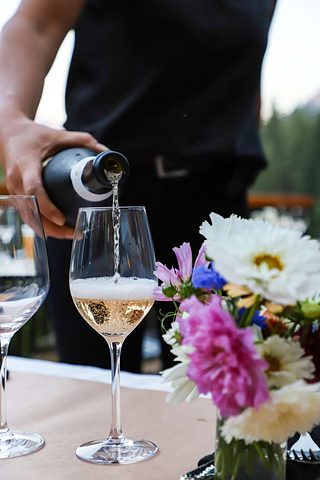 A server pours sparkling wine into a glass beside a colorful bouquet of flowers on a dining table.
